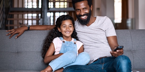 Father and daughter sitting on a couch, father holding a remote